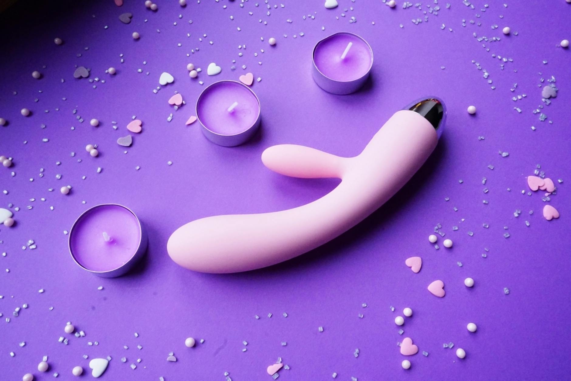 A pink silicone vibrator on a purple surface with heart confetti and candles, representing intimate pleasure.